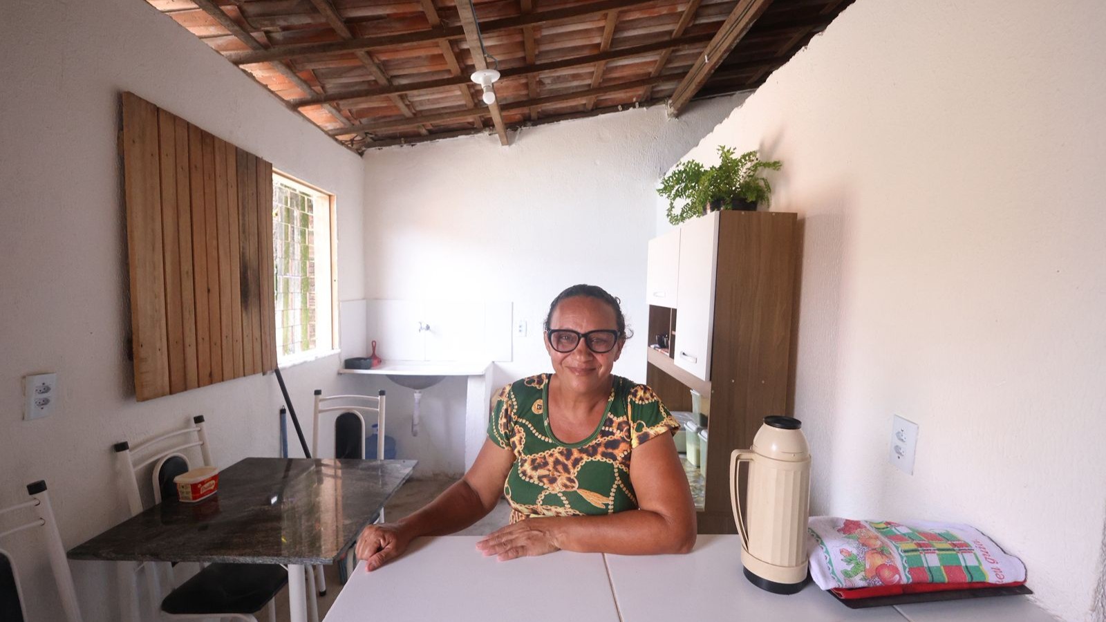 A foto mostra uma mulher de aproximadamente 60 anos sentada à mesa de uma casa bem arrumada, ela olha para a câmera e sorri